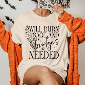 May include: A white t-shirt with black text that reads "Will burn sage and bridges as needed". The text is stylized with a decorative design of a burning sage bundle.