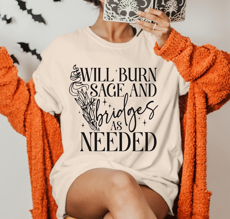May include: A white t-shirt with black text that reads "Will burn sage and bridges as needed". The text is stylized with a decorative design of a burning sage bundle.