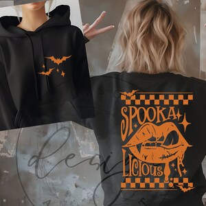 May include: Black hoodie with orange bat and star graphics on the front. The back of the hoodie features the words "Spooka Licious" with a vampire mouth graphic, all in orange. A Halloween-themed sweatshirt.