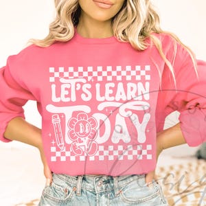 May include: Pink sweatshirt with the text "LET'S LEARN TODAY" in white, featuring a flower with a smiling face and a pencil graphic. The design includes a white and pink checkered pattern.