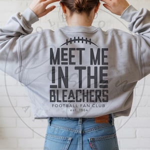 Könnte beinhalten: Hellgraues Sweatshirt mit schwarzem Text "MEET ME IN THE BLEACHERS", einem Football-Grafik und "FOOTBALL FAN CLUB est. 1964". Das Sweatshirt wird mit einer blauen Jeans kombiniert.