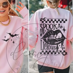 May include: A light pink sweatshirt with black bat and star graphics on the front. The back features the words "Spookalicious" with a vampire mouth graphic, bats, and a checkered pattern. A casual Halloween-themed top.