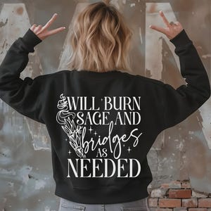 May include: Black sweatshirt with white text that reads "Will burn sage and bridges as needed". The text is accompanied by a stylised image of a burning sage bundle.