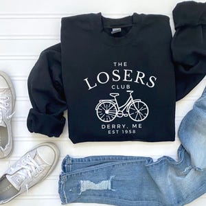 Könnte beinhalten: Schwarzer Pullover mit weißem Text, der "The Losers Club Derry, ME Est 1958" lautet, und einer weißen Fahrradgrafik.