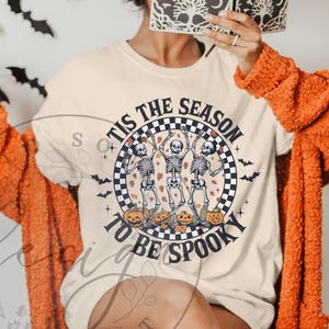 May include: Beige t-shirt with a Halloween design. The shirt features a circular graphic with dancing skeletons, pumpkins, and the text "TIS THE SEASON TO BE SPOOKY". Black bats are scattered around the graphic.