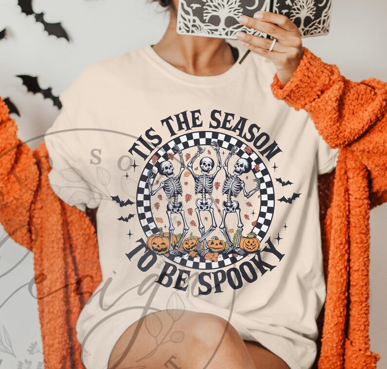 May include: Beige t-shirt with a Halloween design. The shirt features a circular graphic with dancing skeletons, pumpkins, and the text "TIS THE SEASON TO BE SPOOKY". Black bats are scattered around the graphic.