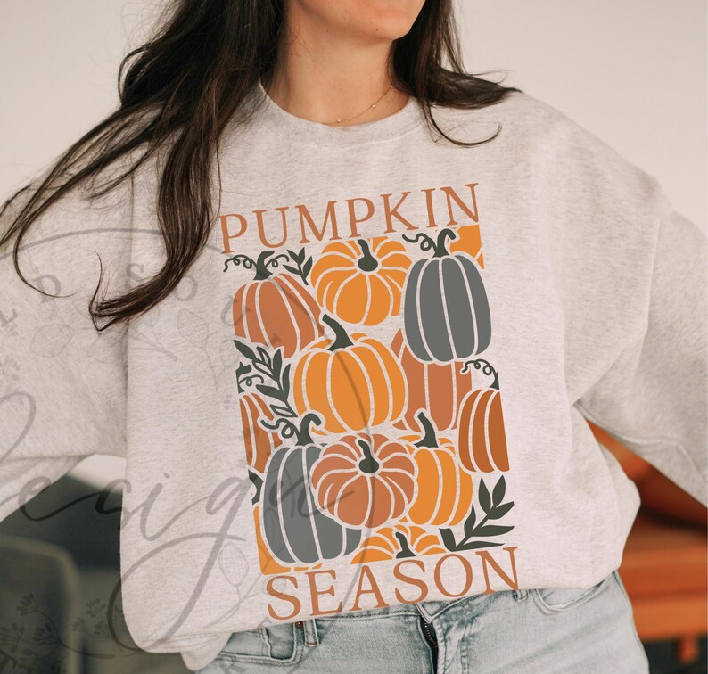May include: A light gray sweatshirt featuring a fall-themed graphic. The design includes orange, brown, and gray pumpkins with the words "PUMPKIN SEASON" in a matching orange color. The sweatshirt is a comfortable, casual style.