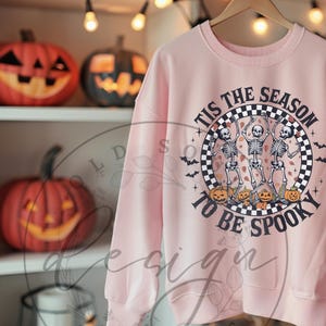 May include: A light pink sweatshirt with a Halloween-themed graphic. The graphic features three dancing skeletons, jack-o'-lanterns, and the text "Tis the Season to be Spooky" in a circular design. Decorative pumpkins are on the shelves.