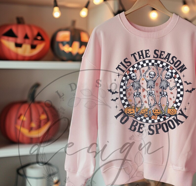 May include: A light pink sweatshirt with a Halloween-themed graphic. The graphic features three dancing skeletons, jack-o'-lanterns, and the text "Tis the Season to be Spooky" in a circular design. Decorative pumpkins are on the shelves.