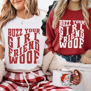 Può includere: Una maglietta bianca e una felpa rossa con la scritta "BUZZ YOUR GIRL FRIEND WOOF" in un carattere audace e stilizzato. L'immagine include anche una tazza bianca con una grafica di Babbo Natale.