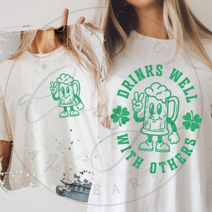 May include: Two t-shirts, one cream and one white, featuring a green cartoon beer mug with a peace sign and the text "DRINKS WELL WITH OTHERS". The design includes shamrocks, perfect for St. Patrick's Day or casual wear.