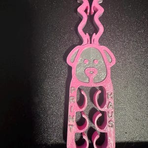 May include: A pink 3D-printed dog-shaped item with the text "PRINT RADS" on the side. The dog has a gray face and curly accents above its head. The item is designed with cut-out shapes.