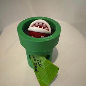 May include: A green 3D-printed pipe with a red and white Piranha Plant from the Super Mario video game series. A green bag with the text "So Doo" is coming out of the pipe.