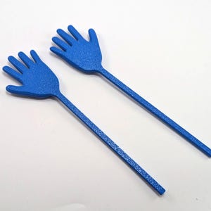 K&ouml;nnte beinhalten: Zwei blaue, handf&ouml;rmige Servierutensilien. Jedes Utensil hat einen langen, d&uuml;nnen Griff und einen handf&ouml;rmigen Kopf mit f&uuml;nf Fingern. Die Utensilien bestehen aus einem strukturierten Material und sind vor einem wei&szlig;en Hintergrund platziert.