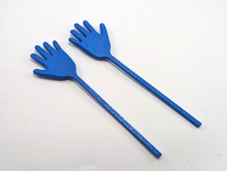 K&ouml;nnte beinhalten: Zwei blaue, handf&ouml;rmige Servierutensilien. Jedes Utensil hat einen langen, d&uuml;nnen Griff und einen handf&ouml;rmigen Kopf mit f&uuml;nf Fingern. Die Utensilien bestehen aus einem strukturierten Material und sind vor einem wei&szlig;en Hintergrund platziert.