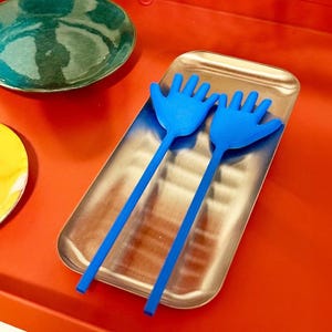 May include: A stainless steel serving tray holds two blue hand-shaped salad servers. A teal and yellow plate are on a red surface. The serving utensils are a unique and playful addition to any kitchen.