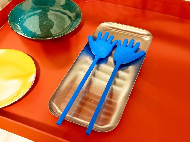 K&ouml;nnte beinhalten: Ein Serviertablett aus Edelstahl enth&auml;lt zwei blaue, handf&ouml;rmige Salatbestecke. Ein teal-farbener und gelber Teller liegen auf einer roten Oberfl&auml;che. Das Servierbesteck ist eine einzigartige und verspielte Erg&auml;nzung.