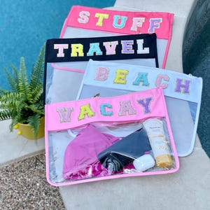 May include: Four clear plastic zippered pouches with different colored fabric fronts. The pouches are labeled "Stuff", "Travel", "Beach", and "Vacay" with colorful letters. The pouch labeled "Vacay" has a pink bikini, a black phone case, and a bottle of sunscreen inside.