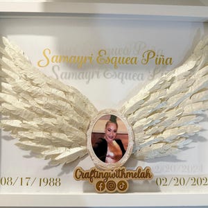 Può includere: Una cornice bianca con sfondo bianco e lettere dorate. La cornice contiene una foto di una donna con ali d'angelo bianche su entrambi i lati. Il testo nella cornice recita "Samayri Esquea Piña" e "Samayri Esquea". Le date "08/17/1988" e "02/20/2024" sono anche incluse nella cornice. La parte inferiore della cornice ha un cartello di legno che recita "Craftingwithmelah" con icone di social media.