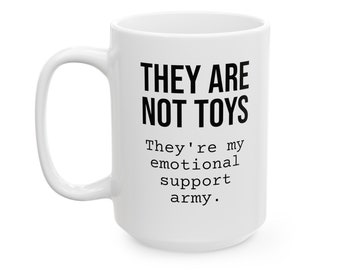 Peut inclure: Mug en céramique blanche avec une grande anse. Le mug porte l'inscription "THEY ARE NOT TOYS" en grosses lettres noires, suivie de "They're my emotional support army." en plus petit.