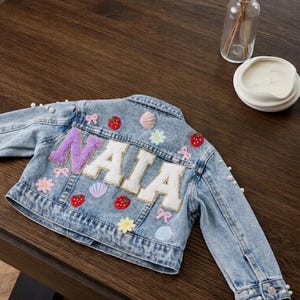 May include: A light blue denim jacket featuring the name NAIA in large, textured letters on the back. The jacket is decorated with embroidered patches of red strawberries, pink bows, colourful seashells, and small flowers, along with white pearl accents.