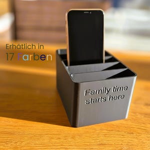 Könnte beinhalten: Ein schwarzer Organizer mit mehreren Fächern, der ein Smartphone hält. Der Text "Family time starts here" ist auf der Vorderseite zu sehen. Das Bild enthält auch den Text "Erhältlich in 17 Farben". Der Organizer steht auf einer Holzoberfläche.