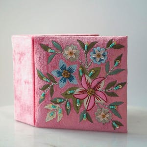 May include: Pink square tissue box cover with embroidered floral design. The flowers are in shades of pink, blue, and white, with green leaves and teal beads. The box is made of a soft, textured material.