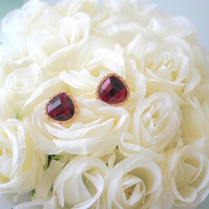 May include: A pair of gold heart-shaped earrings with red gemstone accents. The earrings are displayed on a bed of white roses.
