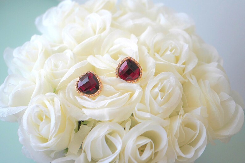 May include: A pair of gold heart-shaped earrings with red gemstone accents. The earrings are displayed on a bed of white roses.