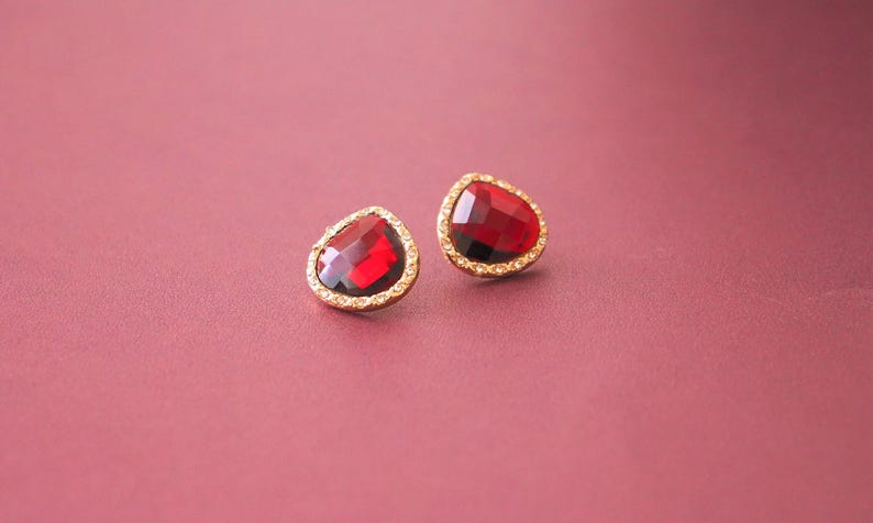 May include: A pair of gold earrings with red gemstone teardrop-shaped stones. The stones are surrounded by a gold setting with small clear stones.