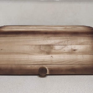 Könnte beinhalten: Eine Brotdose aus Holz mit gewölbtem Deckel und einem kleinen, runden Holzknopf. Die Dose besteht aus hellfarbenem Holz mit dunkleren, verbrannten Kanten. Die Brotdose ist zur Frischhaltung von Brot gedacht.
