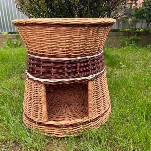 May include: A woven wicker pet bed in natural brown. The bed has a round top and a flared base with an opening. A dark brown band with a white stripe accents the middle. The bed is on green grass.
