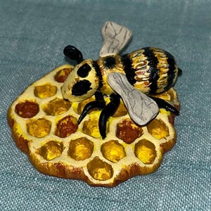 May include: A hand-painted ceramic bee figurine perched on a yellow and brown honeycomb. The bee has black and yellow stripes and white wings.