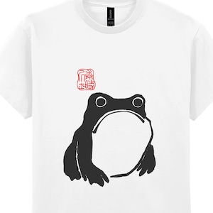 May include: White t-shirt with a black and white illustration of a frog. The frog is facing forward with its mouth open. There is a red stamp above the frog.