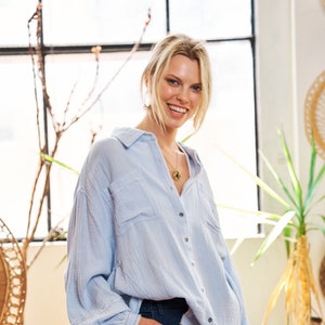 Puede incluir: Una mujer lleva una camisa de manga larga azul claro, de gran tamaño, con botones en la parte delantera y unos vaqueros azul oscuro. Lleva un bolso de crochet blanco en la mano derecha.