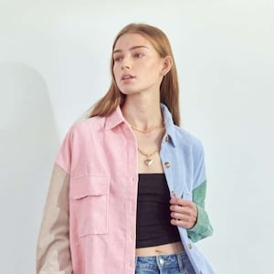 Puede incluir: Una camisa de pana con bloques de color rosa, azul, verde y beige. La camisa tiene dos bolsillos delanteros y cierre de botones. La modelo lleva vaqueros azules y un top negro.