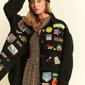 以下が含まれることがあります： 黄色いスマイリーフェイス、「HONEY」、「U.S. ARMY」、「NO BAD VIBES」、「DENIM ARTISTS」などの文字が刺繍されたカラフルなパッチが多数付いた黒のデニムジャケット。茶色のヒョウ柄のドレスと黒のニット帽を着用。