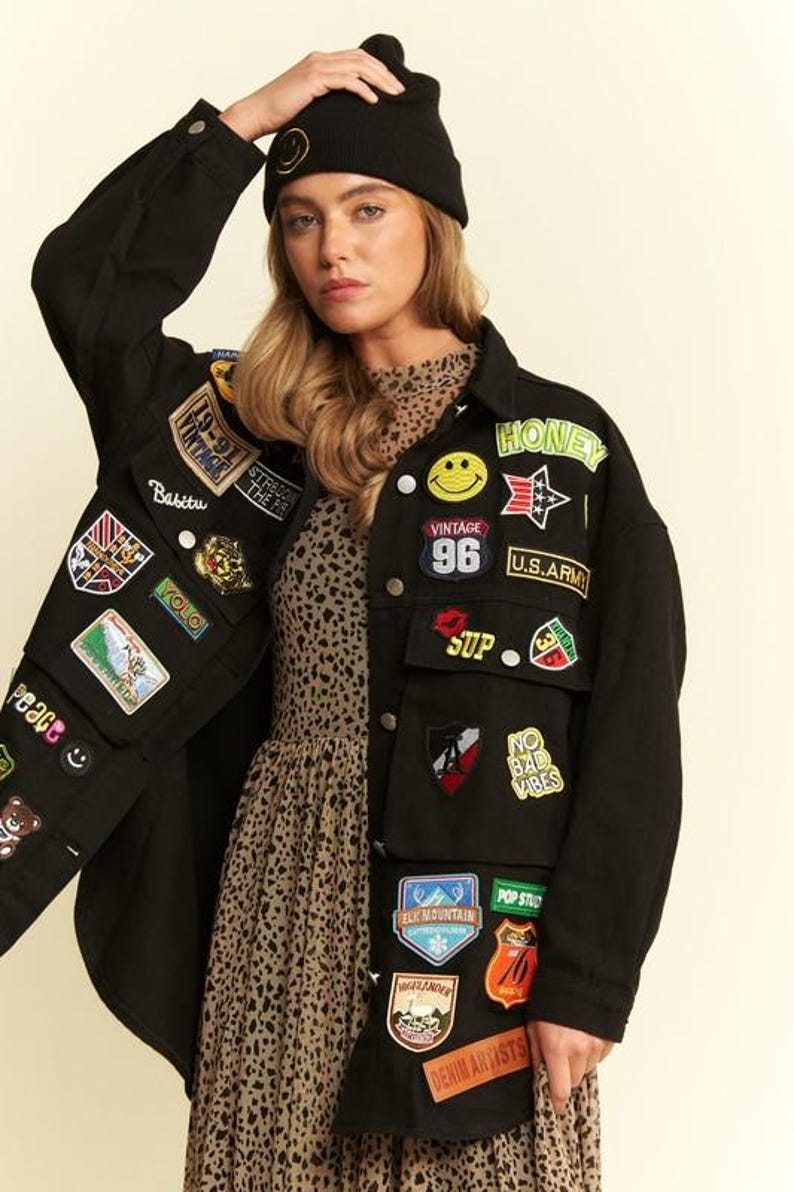 以下が含まれることがあります： 黄色いスマイリーフェイス、「HONEY」、「U.S. ARMY」、「NO BAD VIBES」、「DENIM ARTISTS」などの文字が刺繍されたカラフルなパッチが多数付いた黒のデニムジャケット。茶色のヒョウ柄のドレスと黒のニット帽を着用。