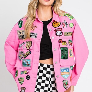 May include: A bright pink denim jacket featuring a variety of colourful embroidered patches, including a smiley face, a teddy bear, and various text-based designs. The jacket has a classic collar, silver-tone metal buttons, and two chest pockets.