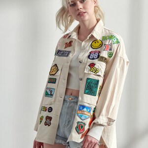 以下が含まれることがあります： 黄色いスマイリーフェイス、HONEYの文字、クマ、その他のグラフィックエンブレムなど、多数のカラフルな刺繍ワッペンで飾られたオフホワイトのボタンダウンジャケット。襟、長袖、フロントポケットが付いています。