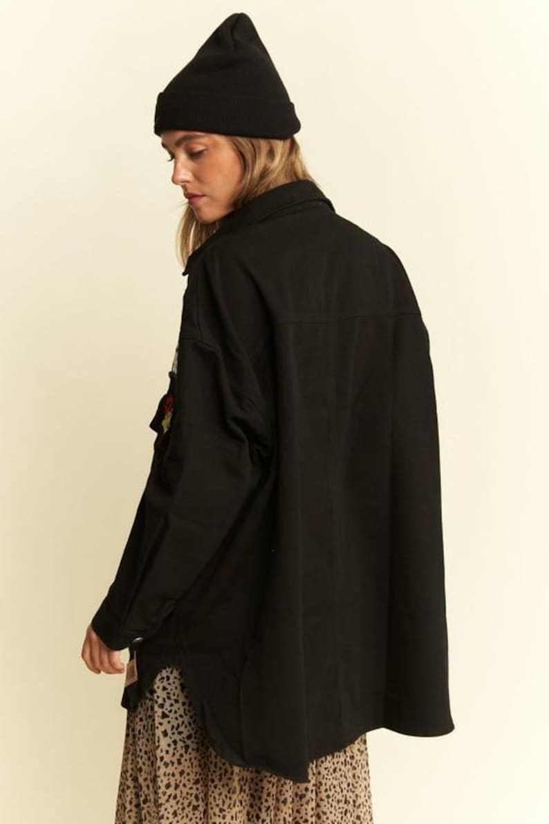 以下が含まれることがあります： 黒いビーニーと、襟と胸ポケットに小さな刺繍パッチが付いた黒い長袖のボタンダウンシャツ。シャツは裾がカーブしたデザインで、黒いヒョウ柄のタンカラーのスカートの上に着用されています。
