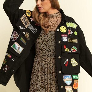 以下が含まれることがあります： 19-91 Vintage、U.S. Army、No Bad Vibesなどのテキストを含む、多数のカラフルな刺繍パッチで飾られた黒の長袖ボタンダウンジャケット。ベージュと黒のヒョウ柄のドレスの上に、黒のニット帽を合わせて着用しています。