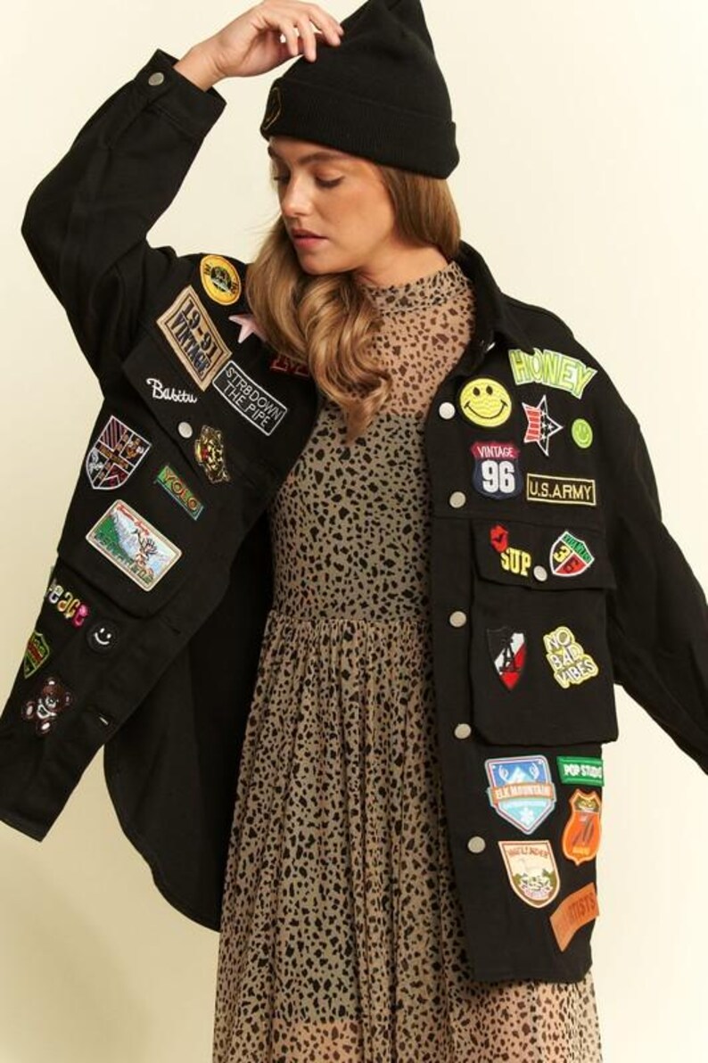 以下が含まれることがあります： 19-91 Vintage、U.S. Army、No Bad Vibesなどのテキストを含む、多数のカラフルな刺繍パッチで飾られた黒の長袖ボタンダウンジャケット。ベージュと黒のヒョウ柄のドレスの上に、黒のニット帽を合わせて着用しています。