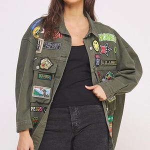 以下が含まれることがあります： HONEY、YOLO、U.S. ARMYなどのテキストを含む、カラフルな刺繍パッチが多数施されたオリーブグリーンのデニムジャケット。黒のタンクトップと黒のデニムショートパンツの上に着用されています。ジャケットはボタン留めで、長袖です。