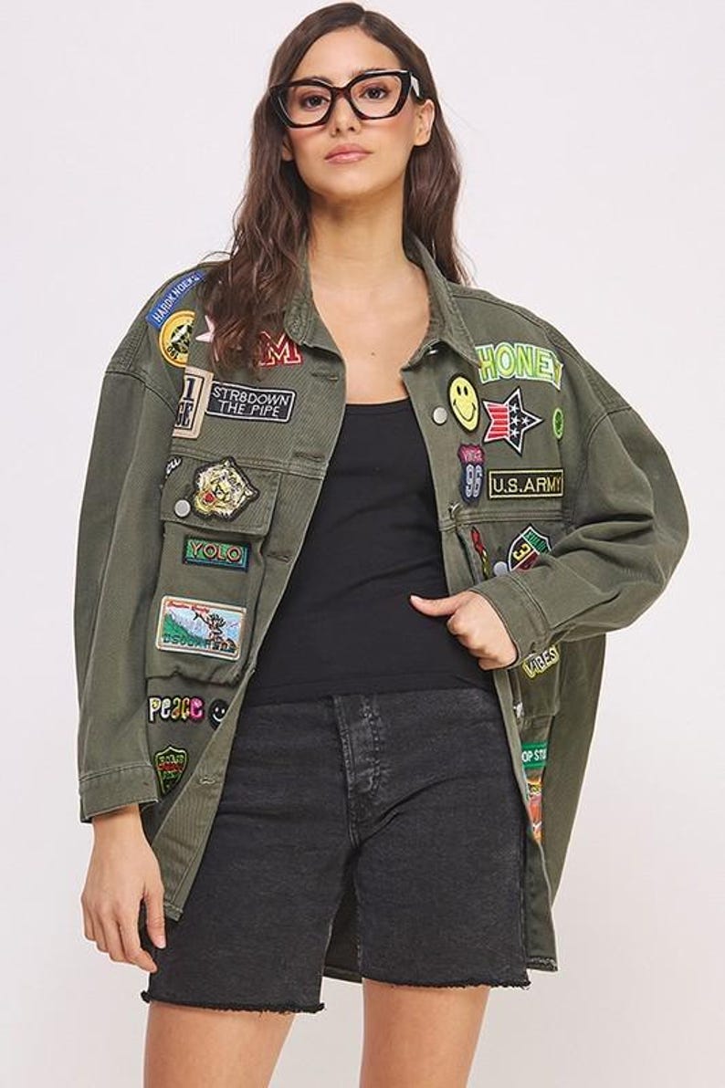 以下が含まれることがあります： HONEY、YOLO、U.S. ARMYなどのテキストを含む、カラフルな刺繍パッチが多数施されたオリーブグリーンのデニムジャケット。黒のタンクトップと黒のデニムショートパンツの上に着用されています。ジャケットはボタン留めで、長袖です。
