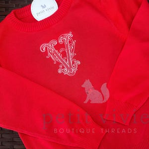 Op de afbeelding: Rode gebreide trui met een witte geborduurde letter V op de voorkant. De trui heeft een ronde hals en lange mouwen. Het label op de trui zegt "petit vivie boutique threads".