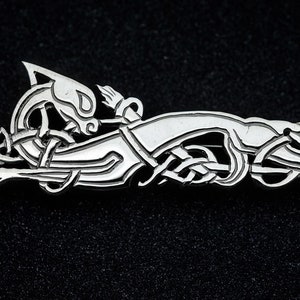 May include: A silver Celtic knot brooch featuring a stylized animal design. The brooch is intricate and detailed, with a knotwork pattern throughout.