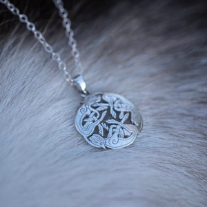 May include: A silver necklace with a circular pendant featuring an intricate design of intertwined animals. The pendant is detailed with a textured surface and a polished finish.