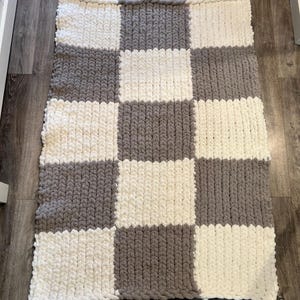 May include: A handmade, chunky knit blanket in a checkerboard pattern. The blanket features alternating squares of soft, off-white and gray yarn. The thick yarn creates a cozy, textured appearance, perfect for adding warmth and style to any space.
