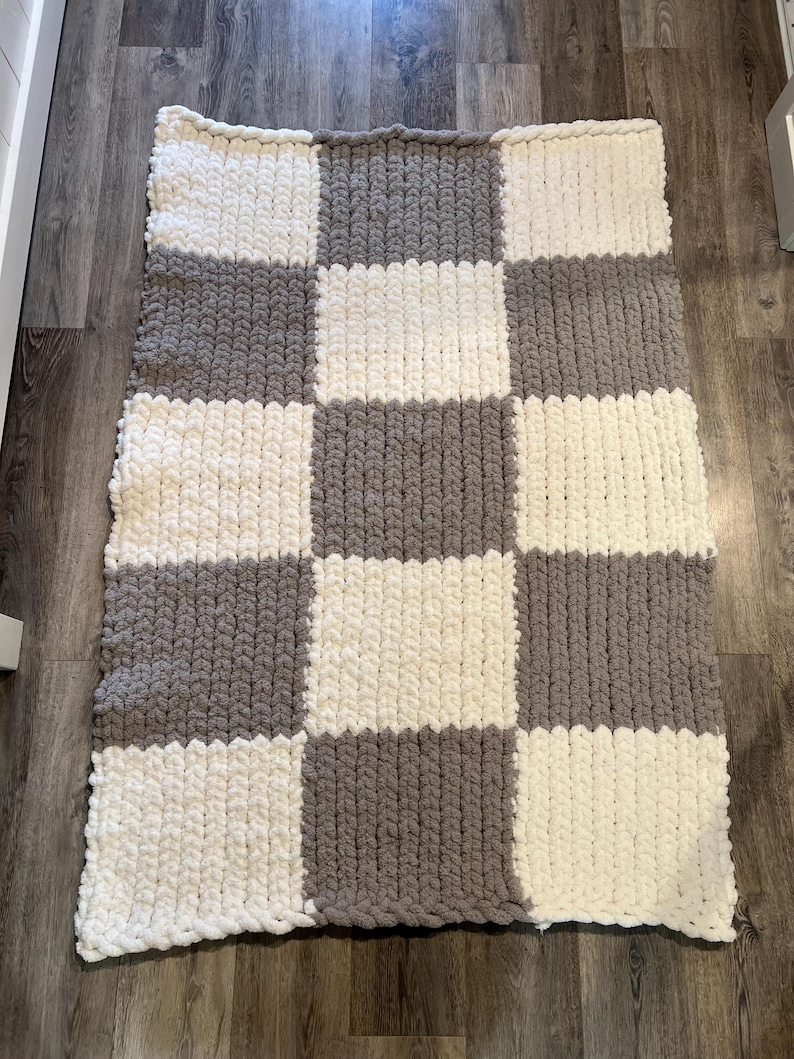 May include: A handmade, chunky knit blanket in a checkerboard pattern. The blanket features alternating squares of soft, off-white and gray yarn. The thick yarn creates a cozy, textured appearance, perfect for adding warmth and style to any space.